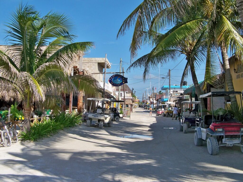 Calle de arena en el pueblo de Holbox con palmeras, carritos de golf y cielo azul