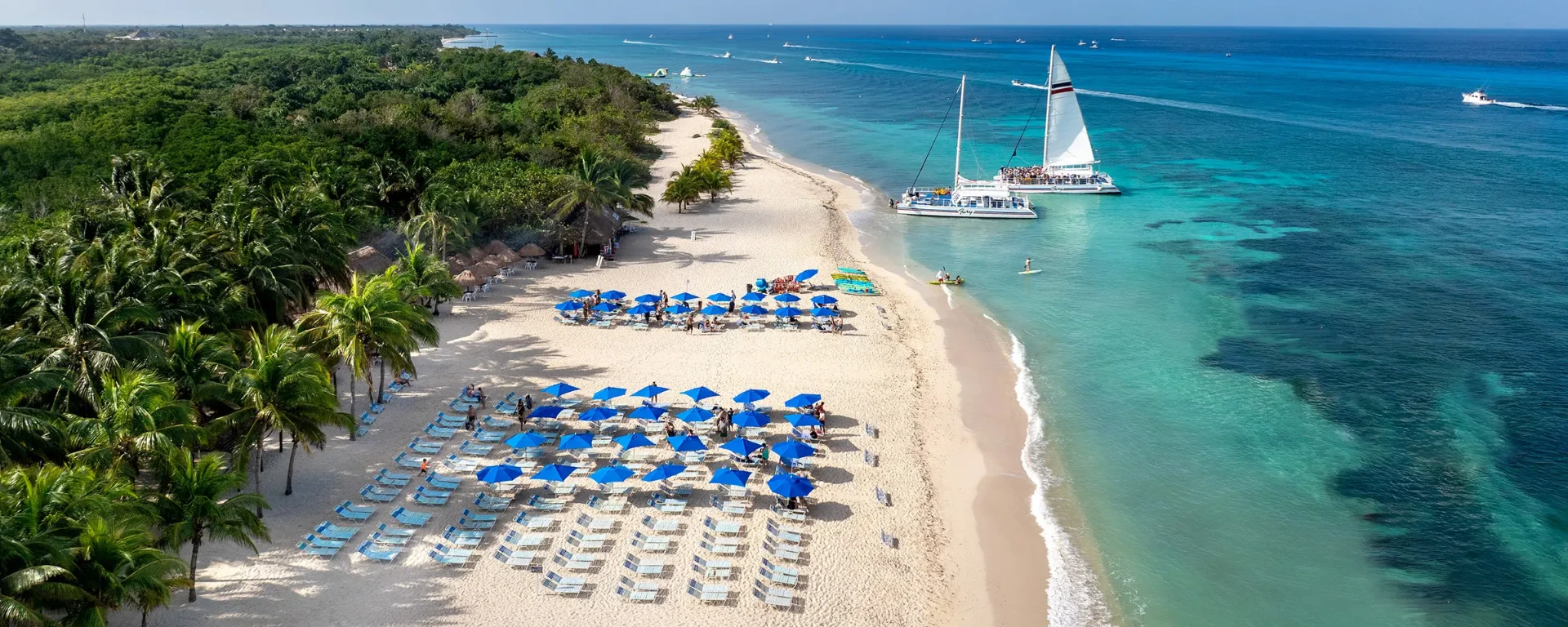 Vista aérea del club de playa de Cozumel con sombrillas azules, catamarán y mar turquesa